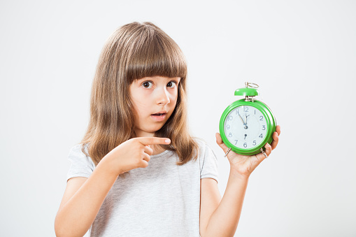 Studio shot portrait of little girl who is pointing at clock that shows five to twelve time. Copy space on gray background.