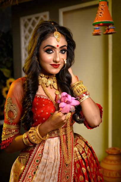 Portrait of pretty young Indian girl wearing traditional saree, gold jewellery and bangles holding flowers in her hands in studio lighting indoor. Indian culture, occasion, religion and fashion. stock photo