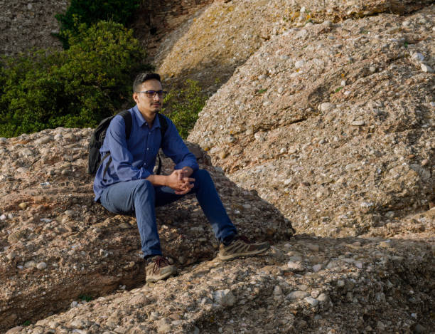 istock Young indian man with backpack sitting on rock looking at nature around himself. 1347202114