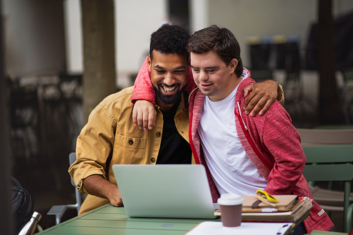 Happy young man with Down syndrome and mentoring friend with arms around outdoors in cafe using laptop