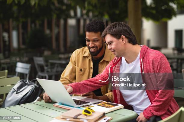 Young Man With Down Syndrome With His Mentoring Friend Sitting Outdoors In Cafe Using Laptop Stock Photo - Download Image Now