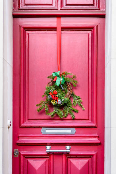 Front view of a Christmas crown made of fir branches, pine cones and ribbons, hanging on the red front door with moldings of a townhouse.