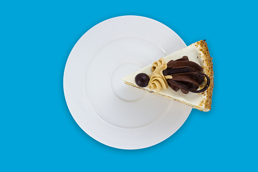 A slice of cake decorated with buttercream and chocolate on a white plate on cyan background. Top view
