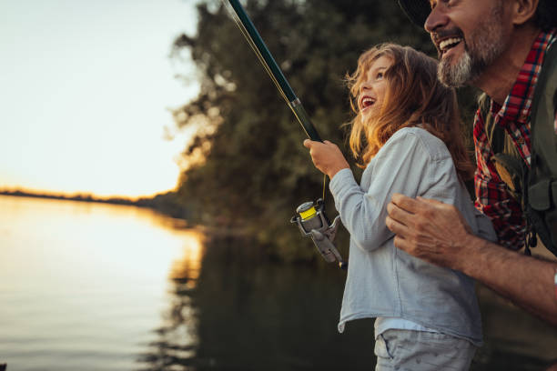 Proud grandfather helping out his granddaughter with fishing Grandfather showing his granddaughter how to fish while standing on the dock by the river. fisher stock pictures, royalty-free photos & images