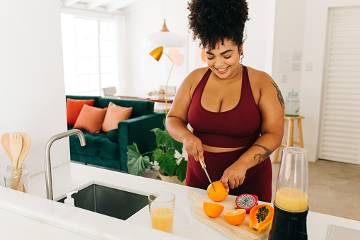 Body positive woman cutting fruits while standing in kitchen. Healthy female in fitness wear preparing juice at home.