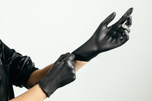 Woman is preparing for her job and putting on black protective gloves. She is wearing black uniform. Photo taken with bright background. She is professional permanent make up artist