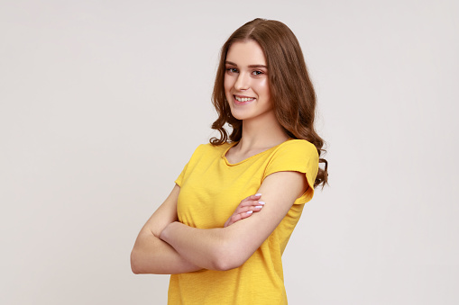 Portrait of positive young woman with brown wavy hair in yellow T-shirt standing with crossed arms, looking at camera with kindness and charming smile. Indoor studio shot isolated on gray background.