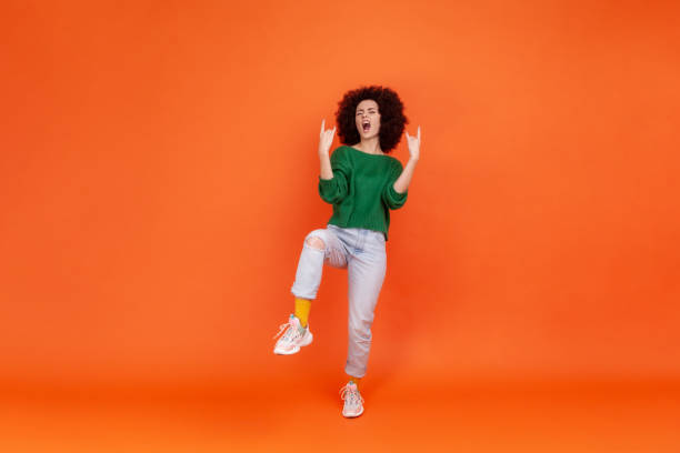 mujer de cuerpo entero con peinado afro con suéter verde de estilo casual que muestra un gesto de rock and roll, de pie sobre una pierna, gritando. - funky people cool women fotografías e imágenes de stock