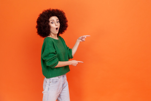 Astonished woman with Afro hairstyle in green sweater pointing aside with finger, having surprised facial expression, keeps mouth open, copy space. Indoor studio shot isolated on orange background.