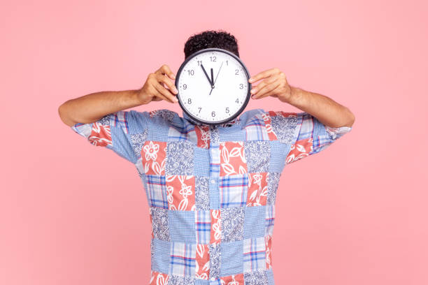 istock Unknown man in blue casual style shirt hiding face behind big wall clock display, wasting his time, procrastination, bad organization of working time. 1347228621