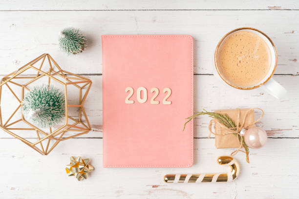 istock Top view Desktop Christmas pink notepad with 2022 letters text. Flat lay on white wooden table background with planner, cup of coffee, candle, toys, Christmas decoration, notebook and stationery 1347236389