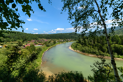 View from the height of the mountain river in the middle of the forest and the village on the shore on a sunny day