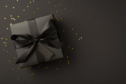 Top view photo of giftbox in black packaging with black satin ribbon bow tag and golden confetti on isolated black background with blank space