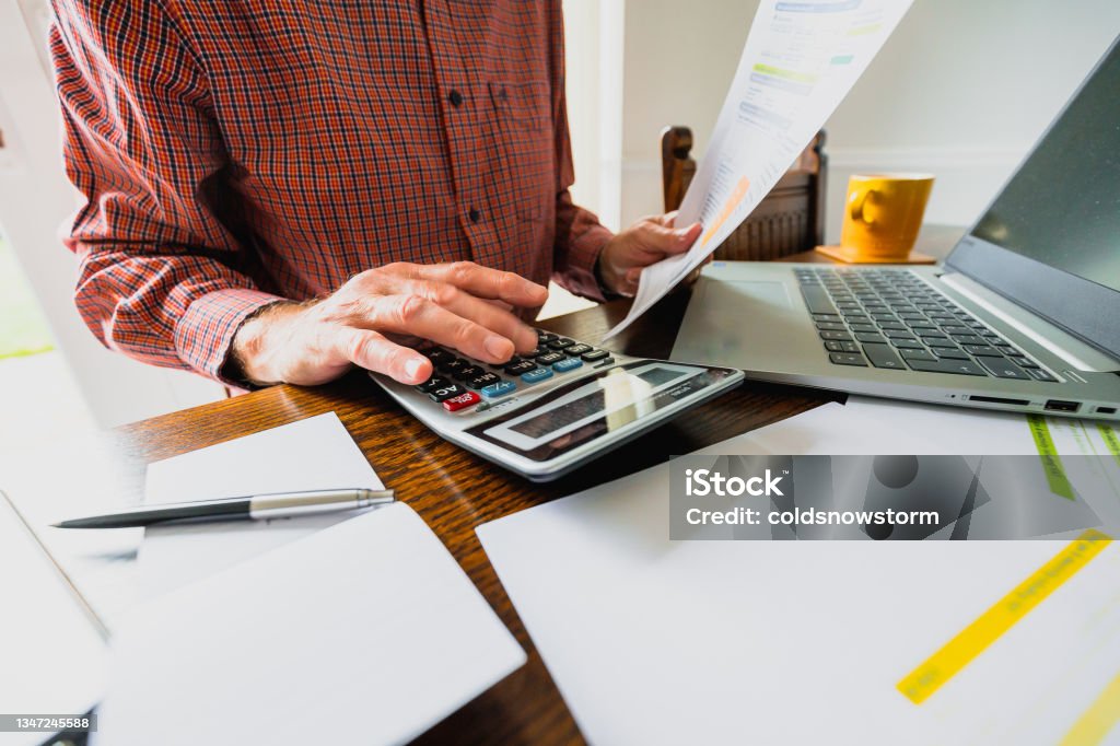 Senior man checking bills at home Wide angle image depicting a senior man's hand holding an energy bill while the other hand checks the numbers on a calculator. The table is strewn with documents and also a laptop. Room for copy space. Financial Bill Stock Photo