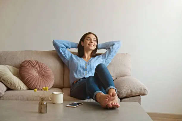 Kick back and relax concept. Young beautiful brunette woman with blissful facial expression alone on the couch with her bare feet on coffee table. Portrait of relaxed female resting at home.