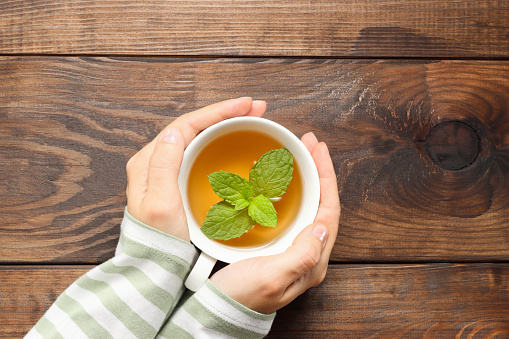 Female hands hold cup of tea with mint leaf. Healthy tea in hands top view on wooden background, copy space