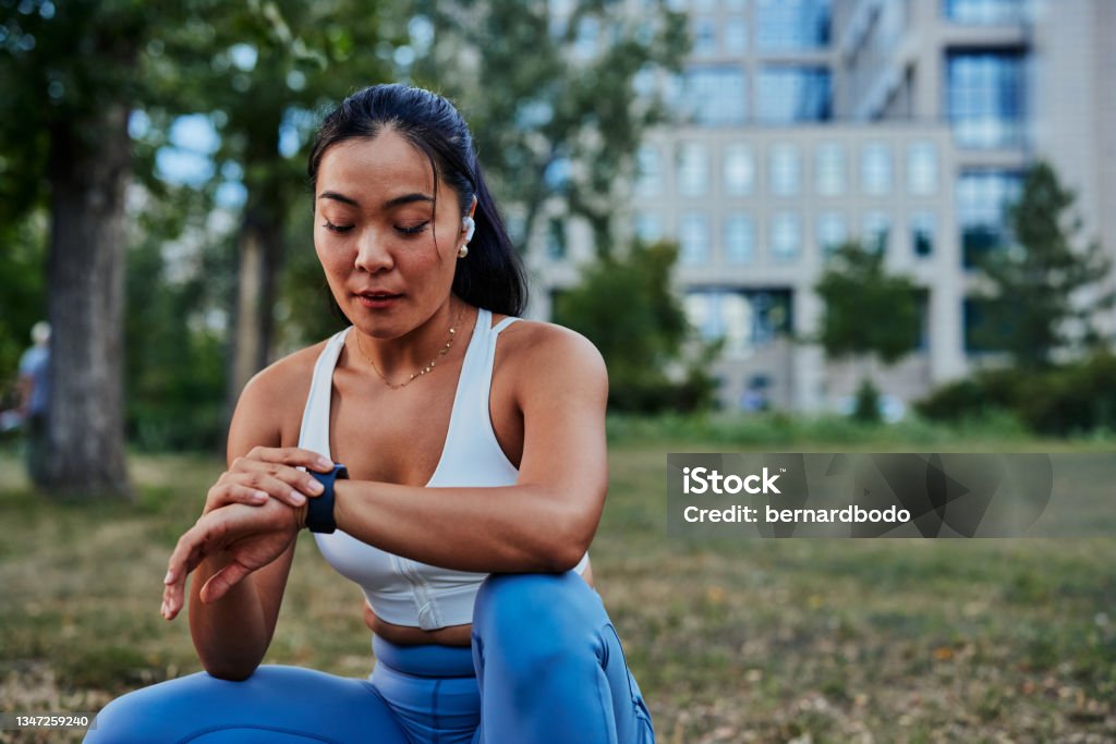 Beautiful sportswoman setting up smart watch during workout in park Female athlete setting up smart watch before workout in a forest. Beautiful asian sportswoman checking data on her gadget outdoor. Exercising Stock Photo