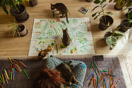 Young female artist, in her cozy room, surrounded with a bunch of plants of different kinds, and her pets, a domestic kitten, drawing a poster of plants