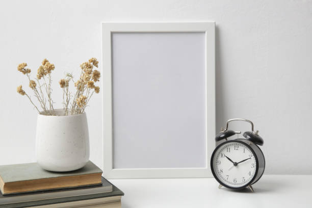 istock Horizontal white frame mockup on table with books, alarm clock and ceramic vase with yellow dry Everlasting flowers. White wall background. 1347269622