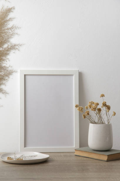 istock White frame mockup on table with books, ceramic vase with yellow dry Everlasting flowers. White wall background. 1347269977