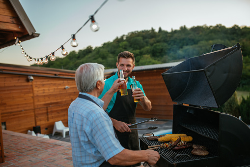 Young man and his elderly father toasting with drinks while having a barbecue in their backyard