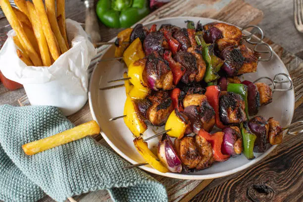 Shashlik with chicken breast, bell peppers and red onions. Served with homemade french fries on a rustic and wooden table. Closeup view from above