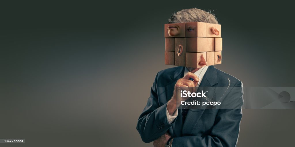 Portrait Of Business Man In Suit With Head As Scrambled Puzzle Pieces A conceptual image of an elderly businessman wearing a dark suit and holding up glasses in contemplation. His head is a jumble of puzzle pieces which are all mixed up, with an eye, ear, mouth etc on each piece. Puzzle Cube Stock Photo