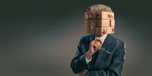 A conceptual image of an elderly businessman wearing a dark suit and holding up glasses in contemplation. His head is a jumble of puzzle pieces which are all mixed up, with an eye, ear, mouth etc on each piece.