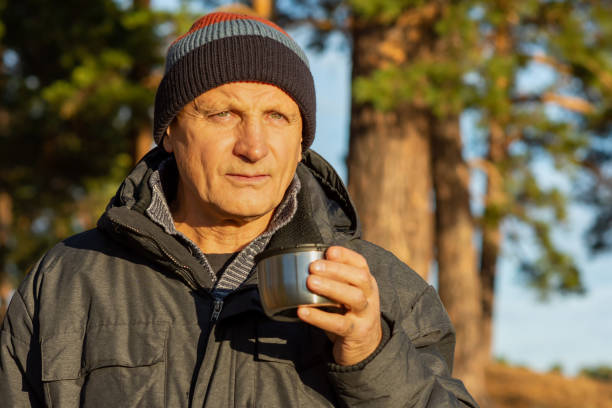 한 노인이 화려한 가을 의 자연을 배경으로 호수 기슭의 보온병에서 뜨거운 차를 마십니다. 자연에서 뜨거운 커피 음료. - russian ethnicity cold relaxation nature 뉴스 사진 이미지