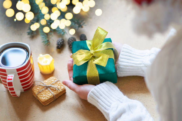 eine frau im weißen pullover hält ein weihnachtsgeschenk auf einem holztischhintergrund. atmosphärisches foto mit bokeh-lichtern. selektiver fokus. konzept der geschenke für weihnachten und neujahr - hot chocolate hot drink high section color image stock-fotos und bilder