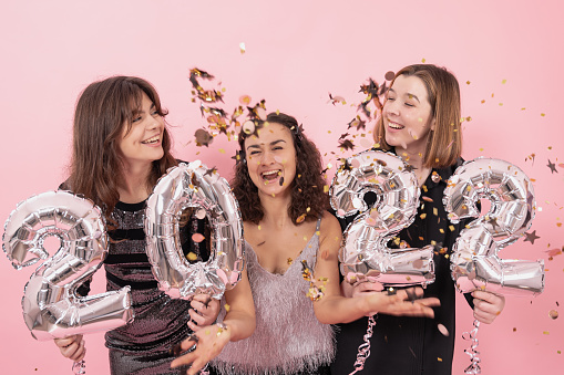 Cheerful girls on a pink background among confetti are holding silver foil balloons with numbers 2022, New Year's fun.