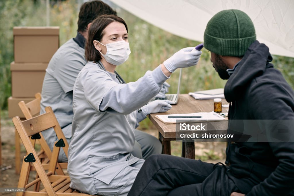 Doctor in protective mask measuring body temperature of sick man Doctor in protective mask measuring body temperature of sick man from migration camp Healthcare And Medicine Stock Photo