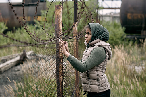 Homeless and lonely girl in casualwear standing by barb wire fence on the territory of refugee camp