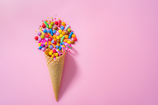Overhead view of a waffle ice cream cone filled with multicolored candies shot on pink background. The composition is at the left of an horizontal frame leaving useful copy space for text and/or logo at the right.\n High resolution 42Mp studio digital capture taken with SONY A7rII and Zeiss Batis 40mm F2.0 CF lens