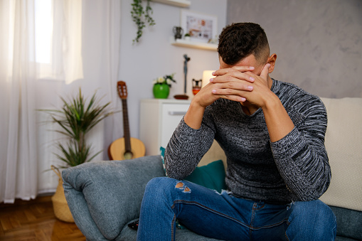Copy space shot of young man sitting on the sofa in his living room, hiding his tears due to anxiety while battling negative thoughts and emotions.