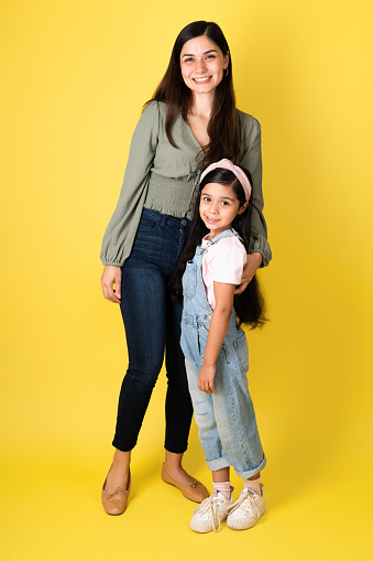 Beautiful young mom and little girl hugging and looking at the camera against a yellow background