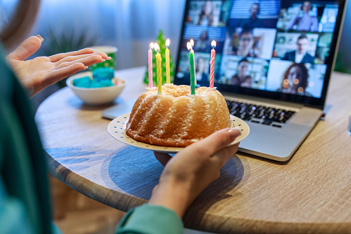 Cut out shot of unrecognizable teenage girl holding a cake with lit birthday candles while her friends, on a video call, are singing ''happy birthday''.