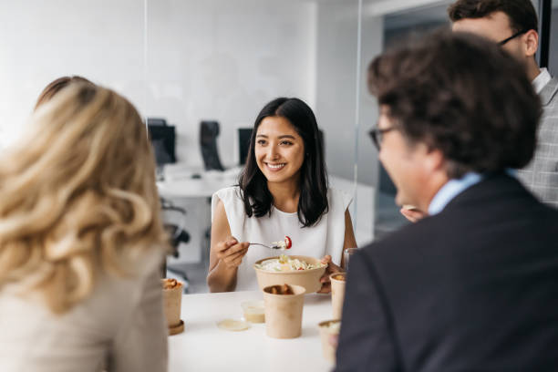 Smiling pretty woman enjoying takeaway lunch at work A good-looking business woman is enjoying a delicious and healthy meal with her colleagues. They are eating in a bright modern office. Horizontal daylight indoor photo. lunch stock pictures, royalty-free photos & images