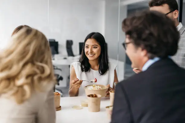 A good-looking business woman is enjoying a delicious and healthy meal with her colleagues. They are eating in a bright modern office. Horizontal daylight indoor photo.