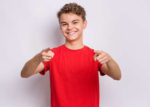 Portrait of happy teen boy pointing fingers at camera. Cute smiling child in red t-shirt choose you, on grey background. Laughing child having fan.