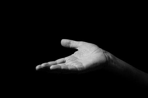 Black & white image of wrinkled male hand held out gesturing, with open palm up, isolated against a black background with dramatic lighting.