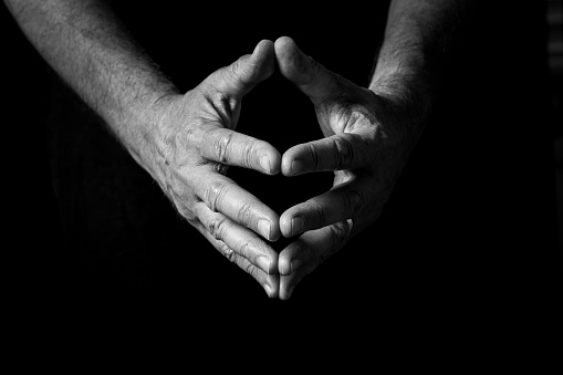 Black & white image of wrinkled male hands held out, clasped with fingers together, in thought or prayer, isolated against a black background with dramatic lighting.