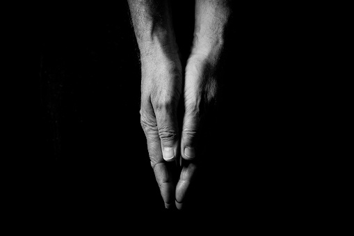 Black & white image of two male hands touching, palms pressed together, isolated against a black background with dramatic lighting.