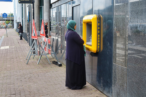 The Hague, Netherlands - August 20 2021 : a muslim woman with head scarf is standing infront of an atm machine on the street