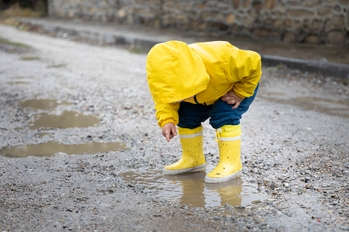 A little cute boy is playing in the street with a puddle. Yellow clothes
