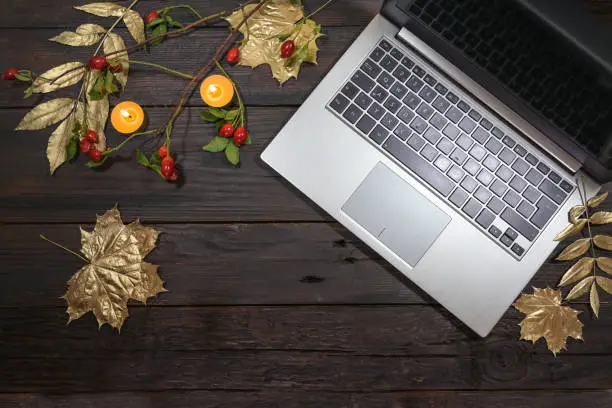 silver laptop computer with gold painted autumn leaves, rose hips and candles on a dark rustic wooden table, seasonal business in the home office, flat lay from above, copy space, selected focus