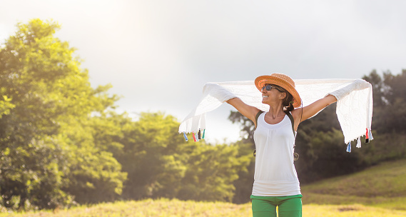 a middle-aged woman with open arms and smiling, holding a white scarf on a sunny day. concept of happiness, fresh air and nature.