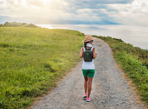 a woman with hat and backpack in the distance, walks along a path towards the sea. in the background a sunset. concept hiking and nature.
