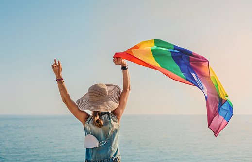 a girl with a hat and her back turned, looks out to sea with a pride flag in her hand.the flag is rainbow and fluttering in the wind.concept of freedom and tolerance. lgtb.