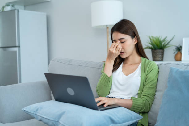Exhausted Asian business woman hurt eye while using laptop computer. Young girl office worker sit on sofa feel visual fatigue and eye strain tired from overwork and massage dry irritable eye at home.